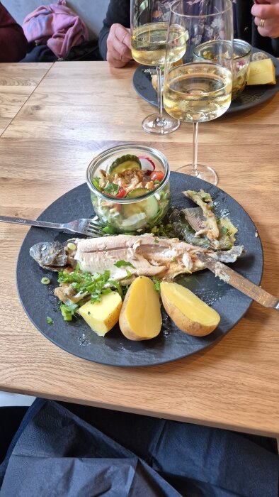 Forell med potatis och sallad på tallrik, två glas vitt vin i bakgrunden.