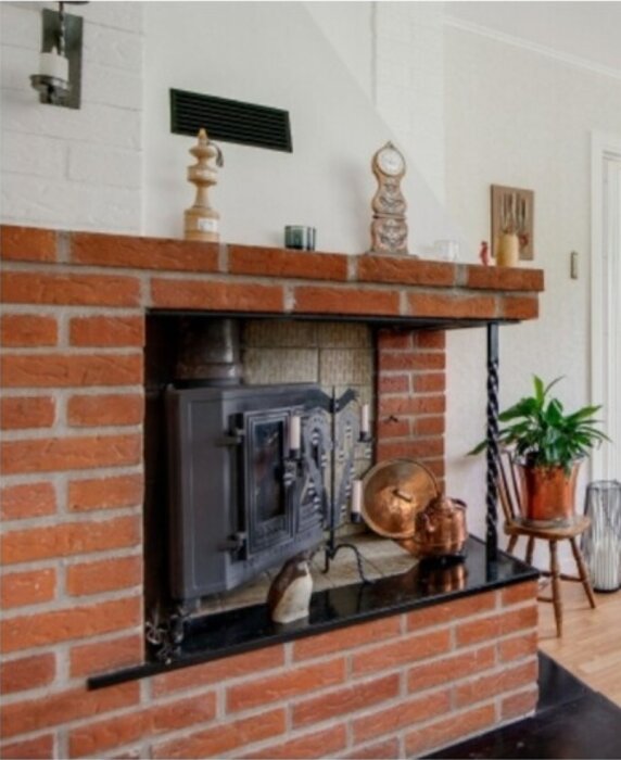 Öppen spis med röda tegelstenar och svart lucka, omgiven av kopparkärl och dekorativa föremål, placerad i ett ljust vardagsrum.