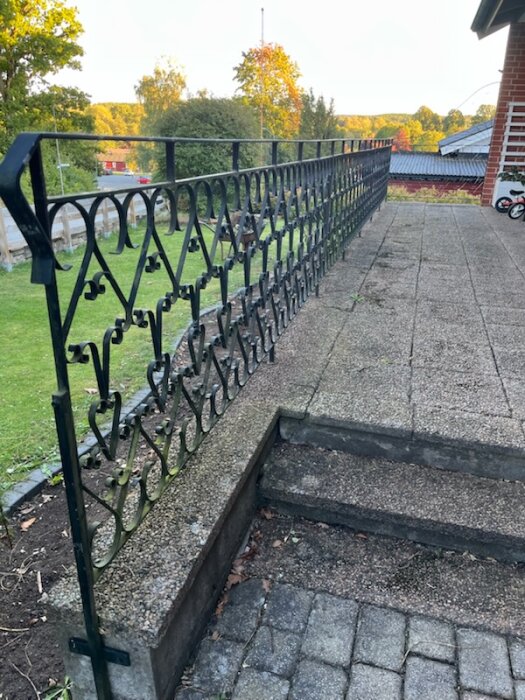 Smidesräcke på altan med dekorativt mönster, sätts fast i plattor och har skruvar i ändarna. Omgivet av grönska och en byggnad i bakgrunden.