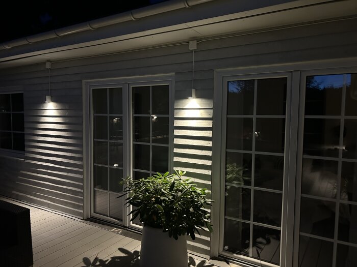 Utomhusbelysning monterad på fasaden vid en altandörr, med ljus som lyser ner och reflekteras på träpanelen och en växt i förgrunden.