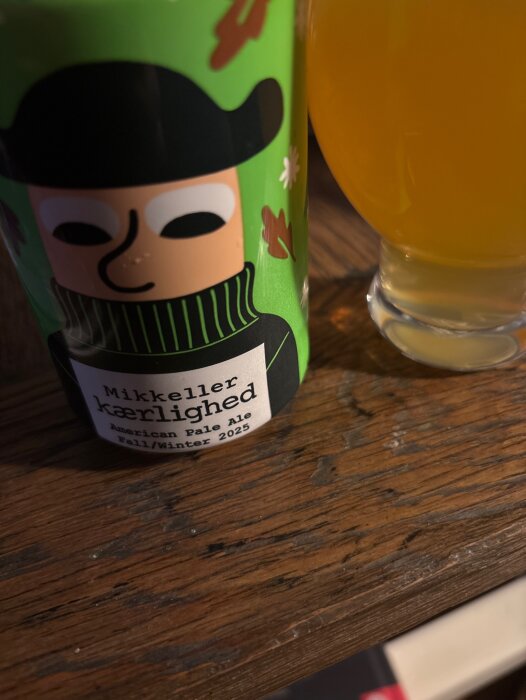 En burk Mikkeller Kærlighed American Pale Ale 2025 och ett glas öl på ett träbord.