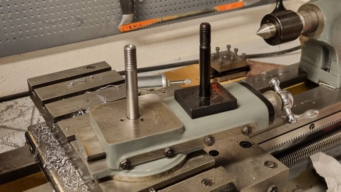 Metallsvarv med två vertikala skruvar; svart platta och svarvad del synliga. Bakgrund med verktyg och spånor ligger på släde.