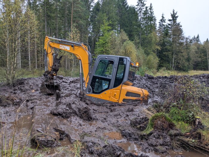 Gul grävmaskin fast i lerig mark vid skogskant, en del av solcellsprojektet.