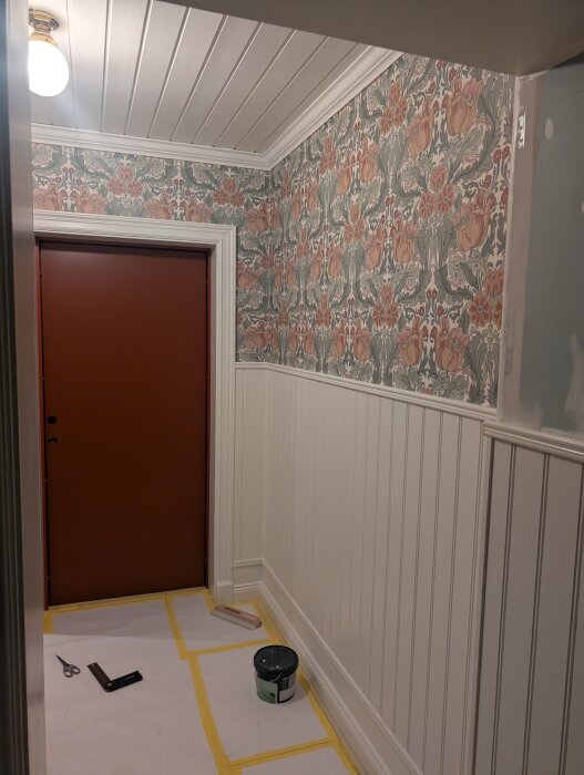 Groventré med ny tapet i blommigt mönster, vitmålad pärlspont på nedre vägg, och tak med panel. Verktyg och färg på golvet.
