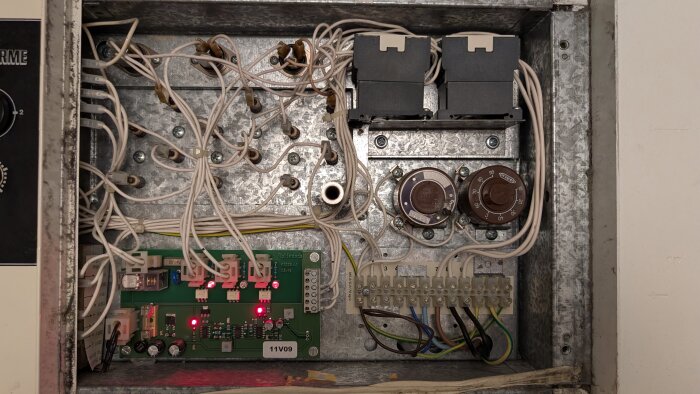 Elskåp med kretskort, kopplingsplintar och termostater, flera kablar anslutna, lysdioder är tända på kretskortet.
