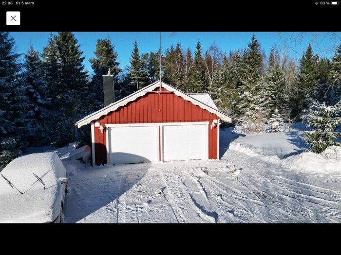 Rött garage med två vita portar omgivet av snö och skog, med snöbelagd infart och parkerad bil framför.