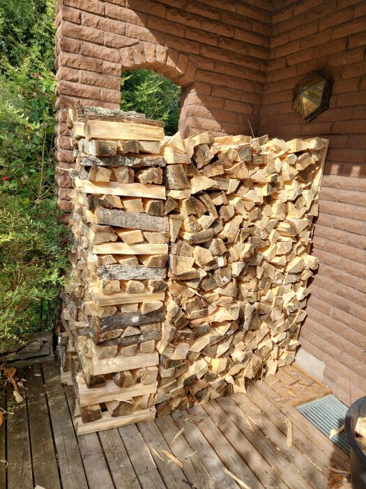 Staplat vedförråd på en träveranda intill en röd tegelvägg, med växter i bakgrunden och en väggklocka ovanför veden.