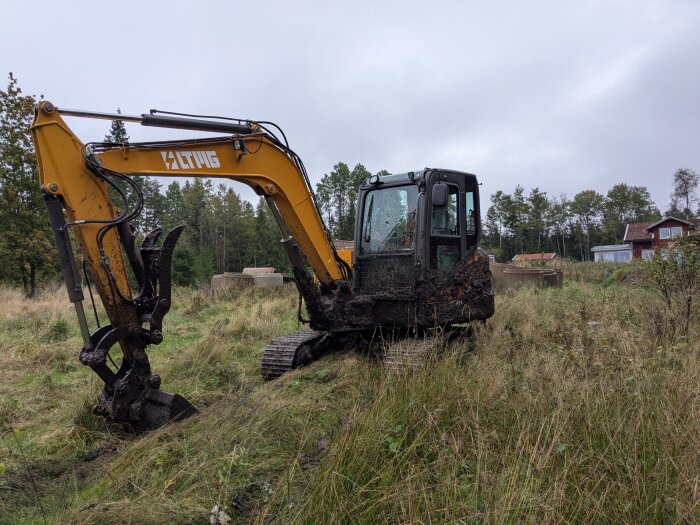 Grävskopa täckt av lera står på en lerig äng vid ett hus, omgiven av träd och grästäckt mark.