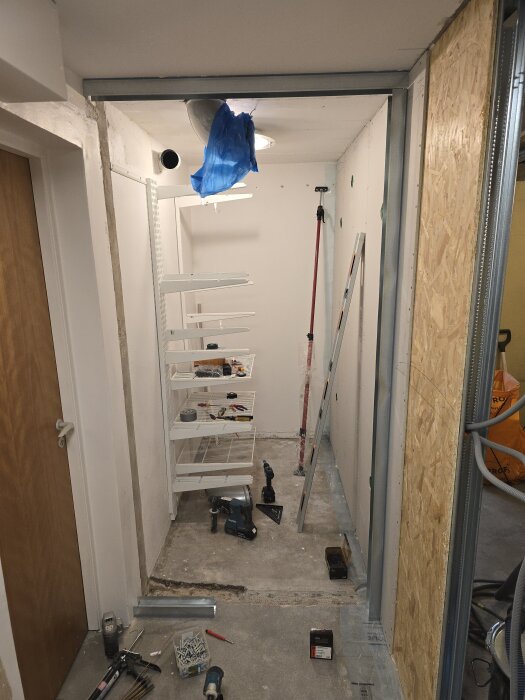 Renoveringsprojekt av klädkammare: vertikala skenor, verktyg, byggmaterial och en stege är framme. OSB-skivor och gipsskivor finns längs väggarna.