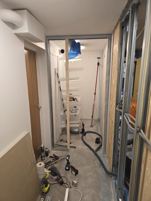 Byggarbetsplats för klädkammare med verktyg, stålskenor, flexrör, betonggolv och en stege; förbereder väggkonstruktion och elektrisk installation.