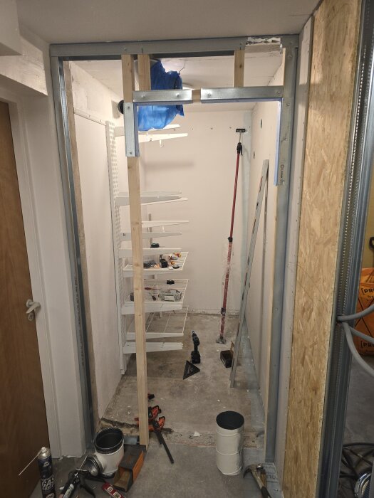 Byggprojekt i klädkammare med stålreglar, träreglar och OSB-skivor. Verktyg och material spridda på golvet. Hyllor och ventilation synliga.