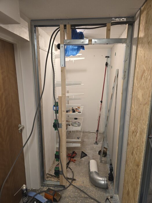 Byggkonstruktion i ett rum med stålskenor och träreglar, flexrör, OSB-skivor och diverse verktyg, under arbete med att bygga en klädkammare.