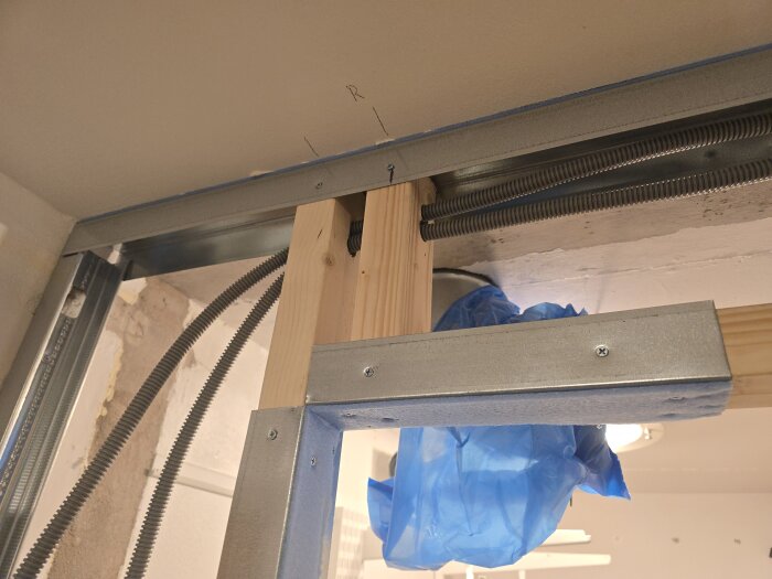 Byggkonstruktion med trä- och ståldelar, flexrör installerade, en brun stående regel och blå plast nära ventilationsrör i dörröppning.