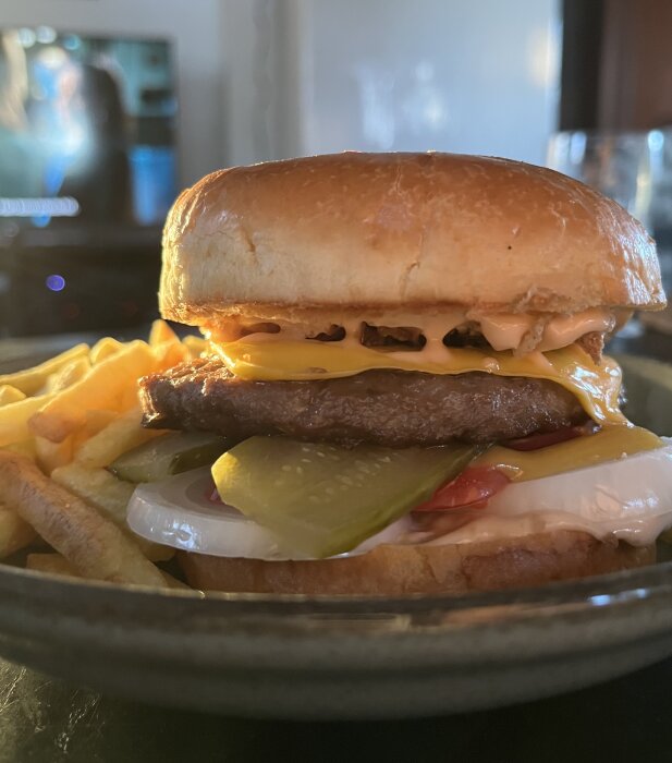 Hamburgare med lök, ost, senap, dressing, rostad lök, tomat, gurka i bröd, serverad med pommes frites på tallrik.
