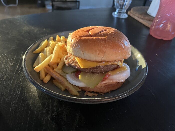 Hamburgare med ost, lök, dressing, tomat och gurka i bröd, serverad med pommes frites på en tallrik.