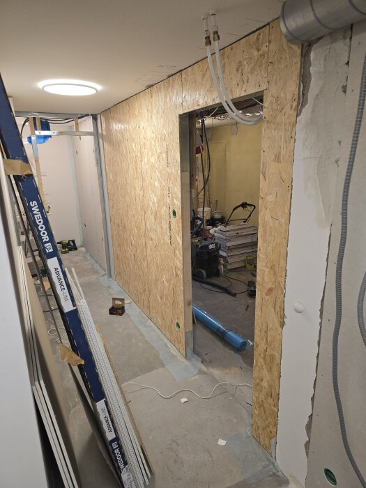 Byggprojekt inomhus med vägg av OSB-skivor, synliga flexrör i taket, och öppning för dörr; golv med verktyg och material på en byggarbetsplats.