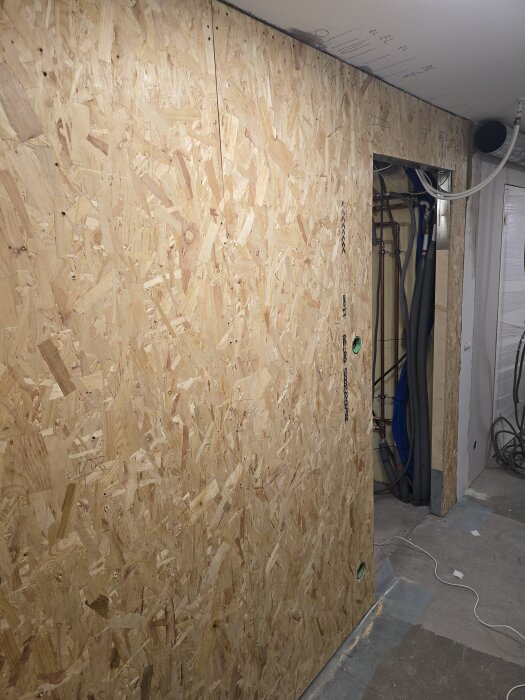 Vägg uppbyggd av OSB-skivor med hål, ledningar och flexrör bakom, samt en dörröppning till vänster i bilden.