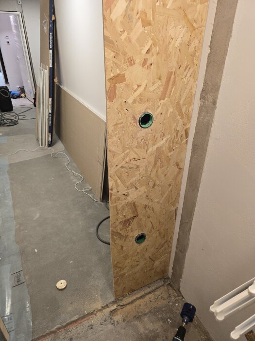 Närbild på en OSB-skiva med två hål för flexrör i en klädkammare under konstruktion, byggmaterial syns i bakgrunden.