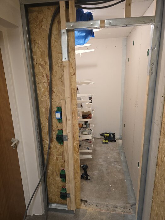 Vertikal skena med träregel i en klädkammare under konstruktion, OSB-skivor och gipsskivor på väggarna, flexrör och verktyg synliga på golvet.