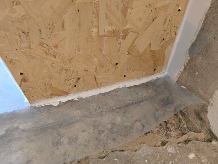OSB-skiva monterad på vägg med vit tätning vid golvet, nära betonggolv och transparent skyddsplast.