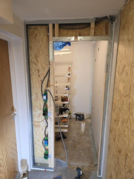 Byggen av en vägg med exponerade elinstallationer och träreglar i en halvfärdig korridor, verktyg och material synliga i bakgrunden.
