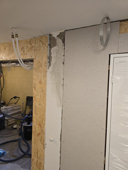 Slutresultat av vägg med monterad gipsskiva och flexrör nära taket, blandade byggmaterial och verktyg i bakgrunden.