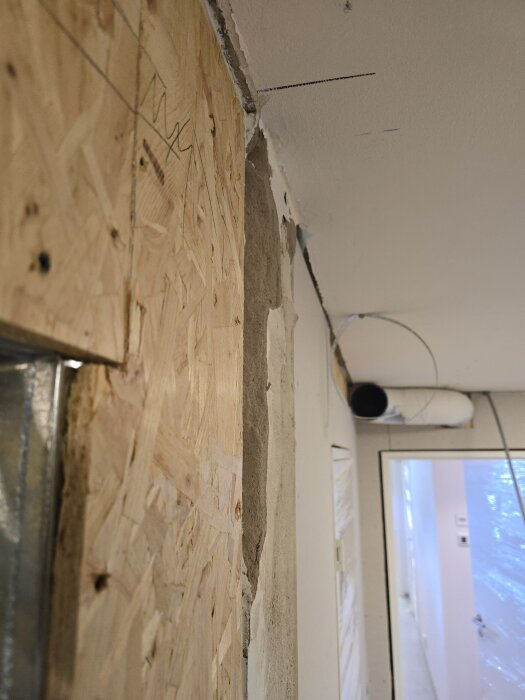 Vägg under konstruktion med OSB-skiva och gipsskiva, synliga ventilationsrör och öppning mot ett förrum täckt med plast.