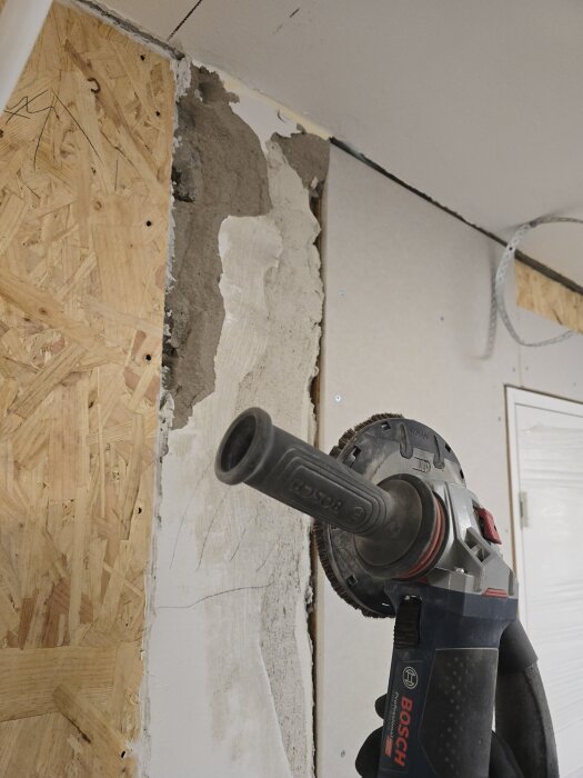 Bild på en handhållen slipmaskin som används för att ta bort puts vid en väggrenovering i ett rum. Bakom slipmaskinen syns osb-skiva och gips.