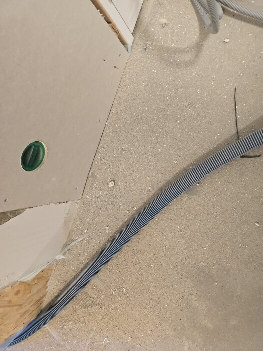 Gipsvägg med hål för eluttag visas, ett kvarvarande flexrör löper parallellt med väggen på ett dammig golv.