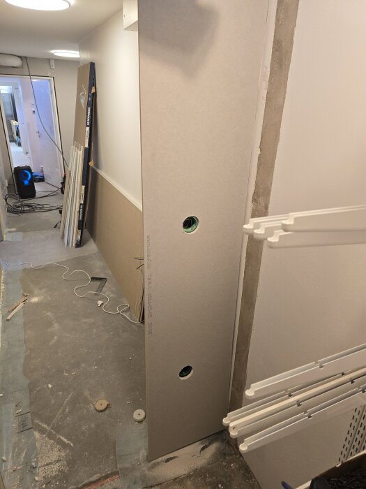 En gipsskiva med hål för eldosor står lutad mot en vägg i en byggarbetsplats med dammigt golv och diverse verktyg.