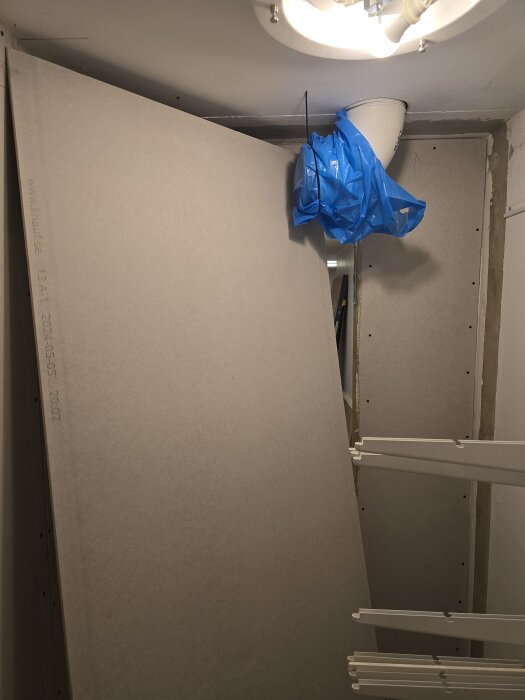 Gipsskiva lutad mot vägg i klädkammare, blå plast på ventilationsrör i taket, inomhusbelysning synlig.