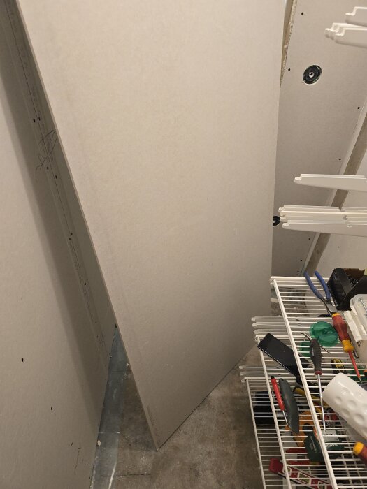 Gipsskiva lutad mot en vägg i klädkammare, verktyg på hylla till höger.