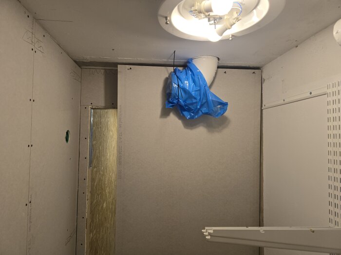 Rum under renovering med gipsskivor på väggarna, en lampa i taket och en fläkt med blå plastöverdrag installerad.