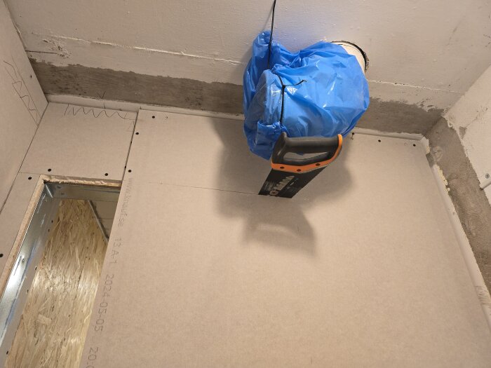 Gipsskiva monterad på vägg med en blå plasttäckning kring en ventilationskanal. En såg med orange handtag hänger nära gipsskivan.