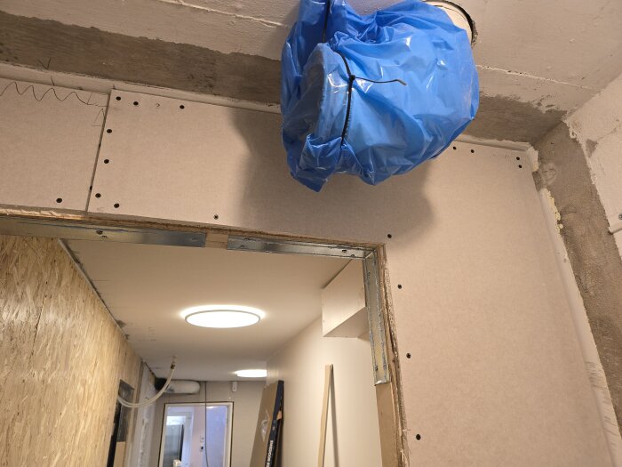 Gipsplattor monterade i en korridor med en luftventilation täckt av blå plast, förberedelse för belysning och arbetet med inredning pågår.