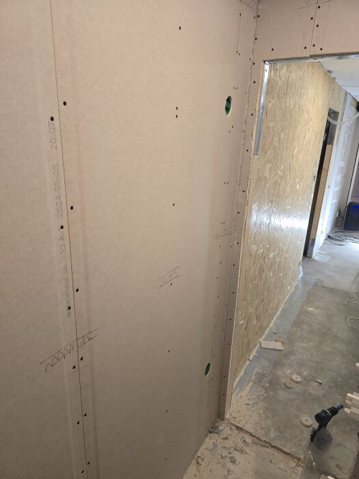 Gipsskivor fastsatta på vägg, med utskuret hål för eldosor och golv täckt av byggdamm. Verktyg och material syns på golvet i korridoren.