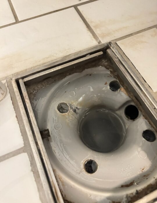 Golvbrunn i ett badrum med synliga små hål och kakelplattor runtomkring, efterfrågar lösning för att undvika dålig lukt och smutsansamling.