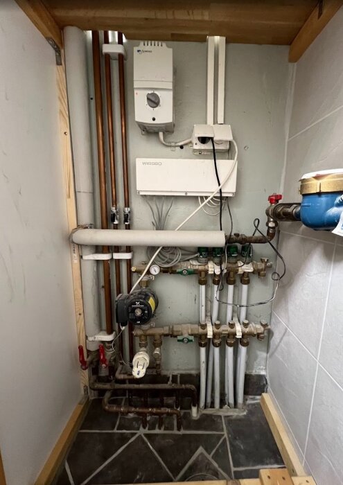 Bild av golvvärmefördelare med shuntgrupp; visar kopparrör, Wirsbo styrbox, cirkulationspump och anslutna rör mot väggen i ett teknikrum.