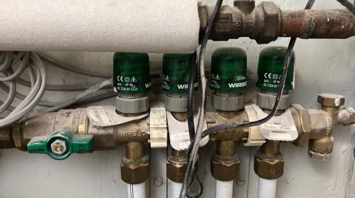 Golvvärmefördelare med fyra gröna styrdon från Wirsbo och vita rör, omgiven av synliga kablar och rörinstallationer.