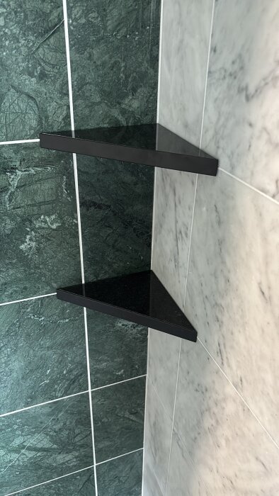 Närbild på kaklat hörn i ett badrum, med två svarta triangulära hyllor mot grön och vit kakelvägg.
