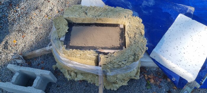 Betongblock omgiven av isolering och frigolit på en grusplan under byggprojekt. Blå presenning och avsågad träbit synlig i närheten.