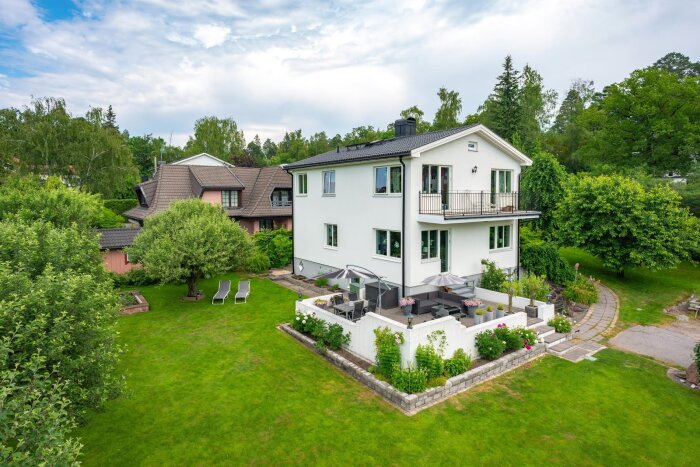 Vit putsad villa från 1935 med två våningar, omgiven av grönska och en trädgård med utemöbler och solstolar.