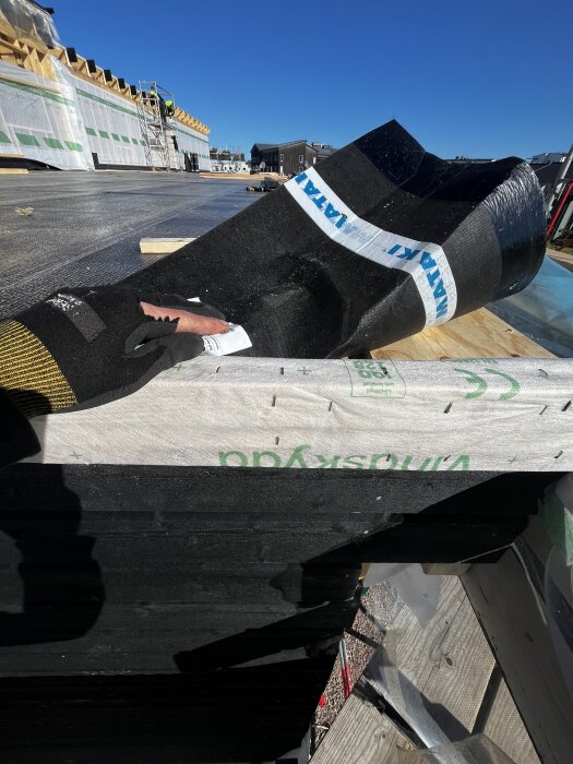 Byggnadstak med monterad vindduk, gloved hand hanterar dukens kant; ställning och arbete pågår i bakgrunden under klar himmel.