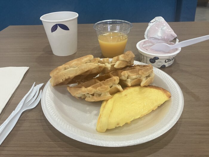 Frukost på ett bord med våfflor, en omelett, apelsinjuice, jordgubbsyoghurt, en tom pappersmugg och plastbestick.
