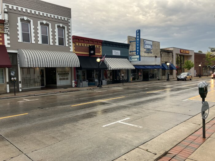Gata med affärer, inklusive möbelaffär och bank, samt parkeringsmätare och bilar under mulen himmel.