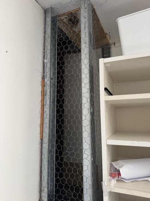 Ett ventilationsrör omgivet av ett hönsnät i ett garage. Grå skiva som verkar ha gått sönder, eventuellt fiberaktigt material synligt.