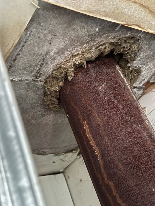 Rostigt ventilationsrör genom vägg med skadad grå skiva och brunt fibermaterial synligt runt röret i garage.