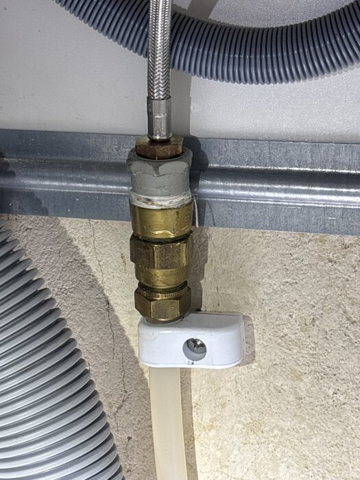 Vit vattentillförselslang ansluten till mässingskoppling under diskbänk med två synliga sexkantsmuttrar, grå flexibla slangar i bakgrunden.