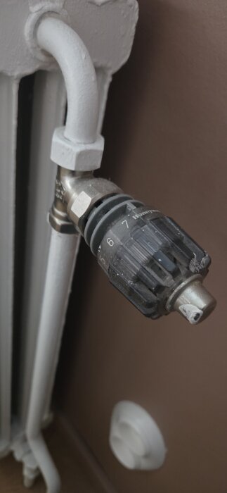 Termostatventil på ett element med inställningsmöjligheter numrerade 0-6, omgiven av en vit rörinstallation och brun vägg i bakgrunden.