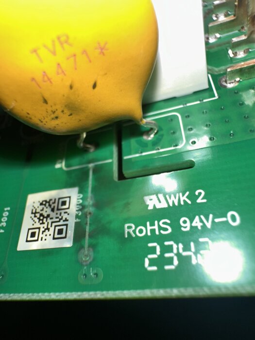 Kretskort med gul komponent (TVR 14471, MOV) och svart bränd fläck nära den, med QR-kod och märkningar på grönt kretskort.